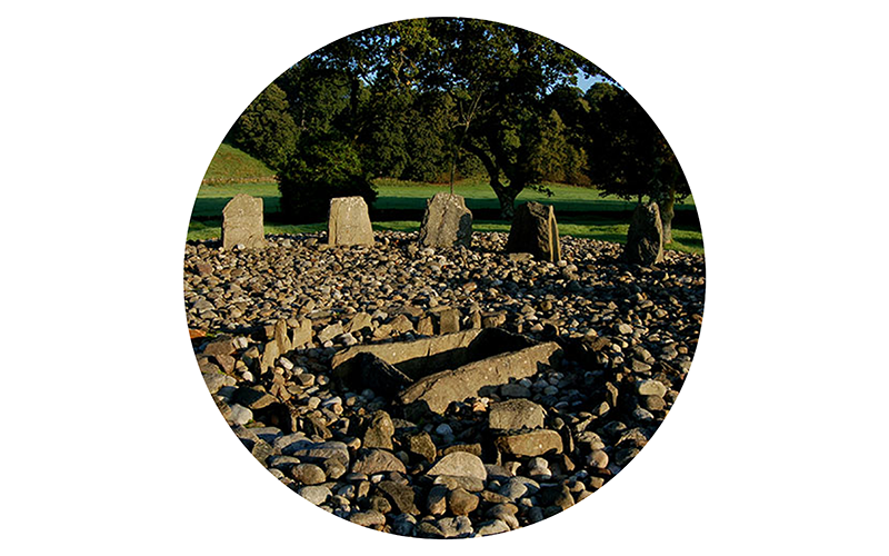 Temple Wood stone circle with large standing stones arranged in a circular pattern, surrounded by a cairn, with trees and grass in the background.