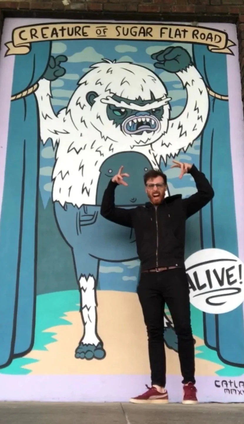 Dave Connis stands in front of a large illustrated mural of a cartoonish yeti, a white ,furry monster with an open mouth and raised arms. Dave mirrors the yet's pose, holding both hands up with fingers spread.