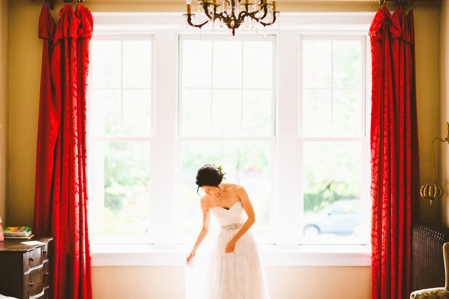 050 - bride adjusting her dress in front of big picture window with red curtains.jpg