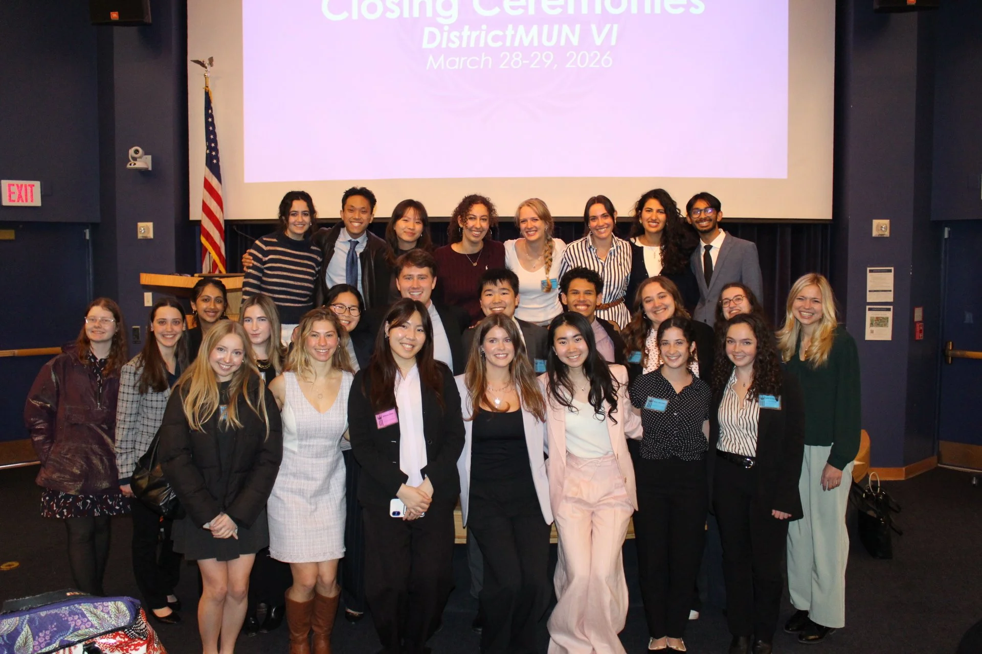 Group of young adults posing together on stage after a closing ceremony at District MUN VI on March 28-29, 2026. They are smiling and dressed in business casual and formal attire, with some wearing name tags. Behind them is a large screen displaying event details, an American flag, and a dark blue wall with exit signs and a security camera.