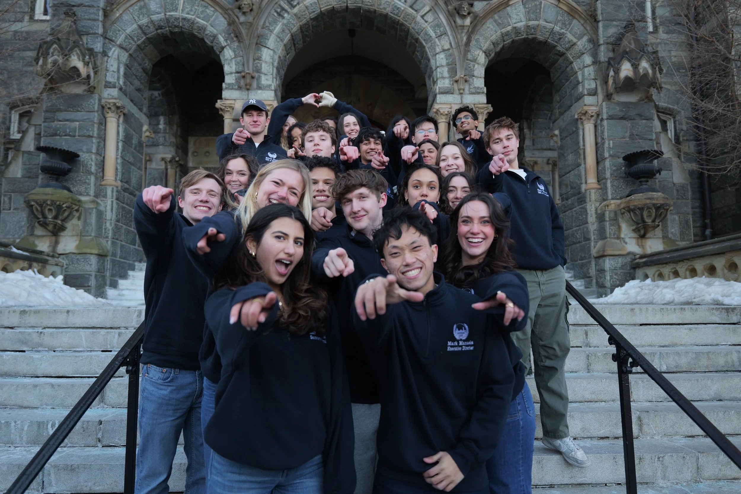 Group of smiling young students standing on the steps of a historic stone building, making heart shapes with their hands, wearing matching navy blue jackets.
