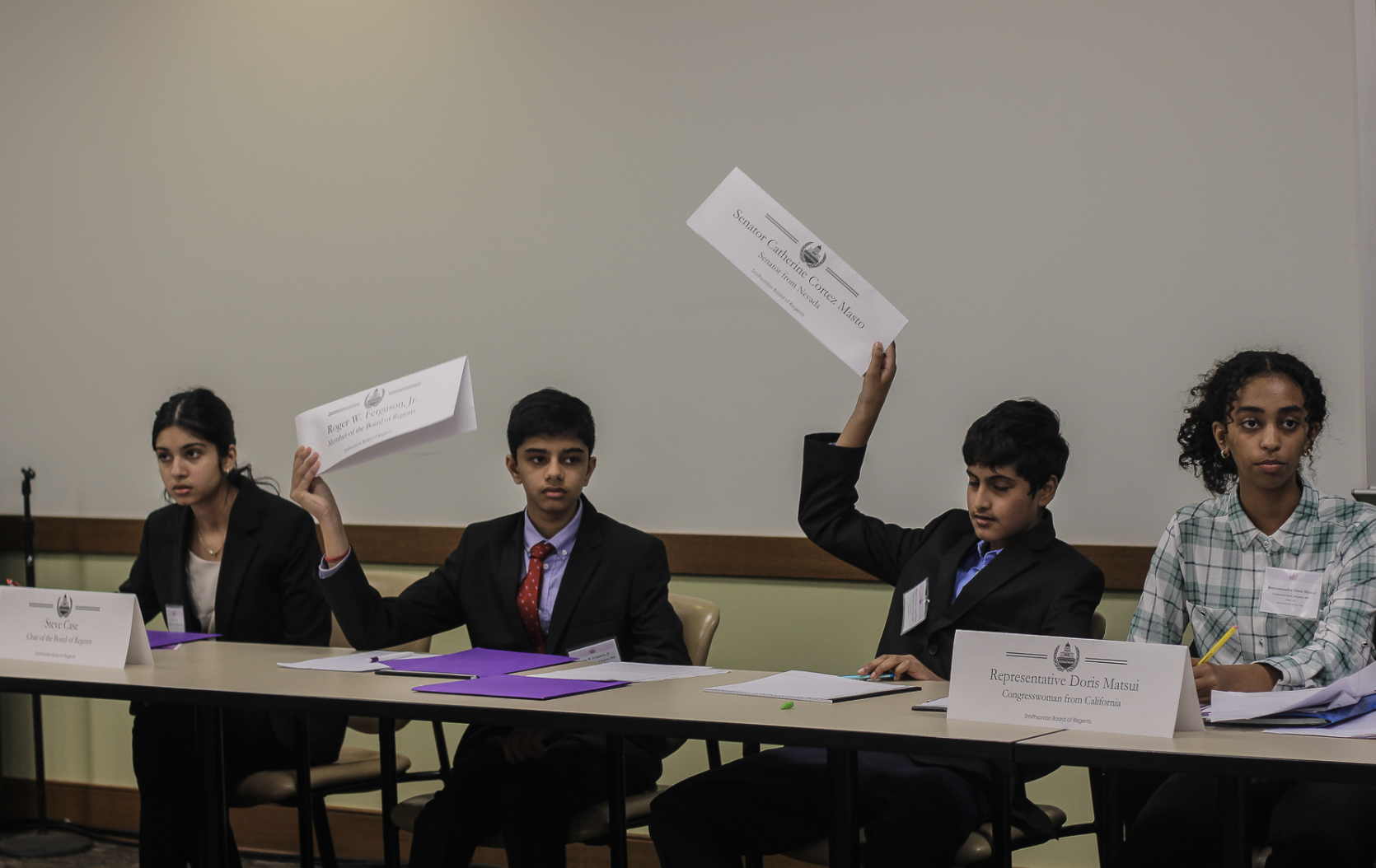 Four children in formal attire participating in a model government or debate, seated at a table with nameplates and purple folders, one boy raising his hand with a placard.