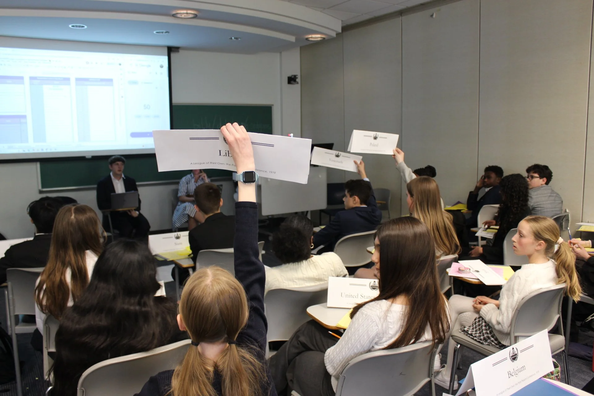 Students in a classroom participating in a Model United Nations conference, holding country name placards such as Belgium and Poland, with a speaker at the front, a projected slide on the screen, and a green chalkboard in the background.
