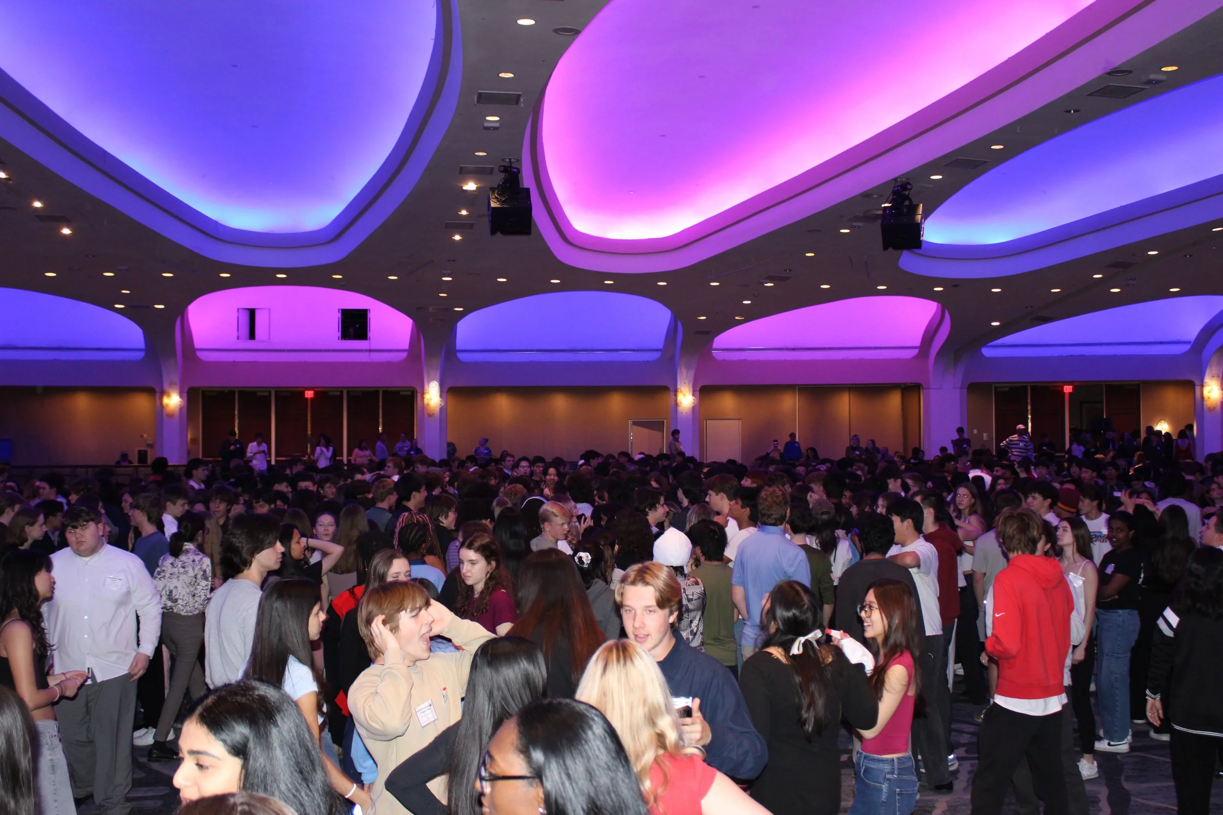 A large group of people gathered in a spacious indoor venue with purple and blue lighting on the ceiling.