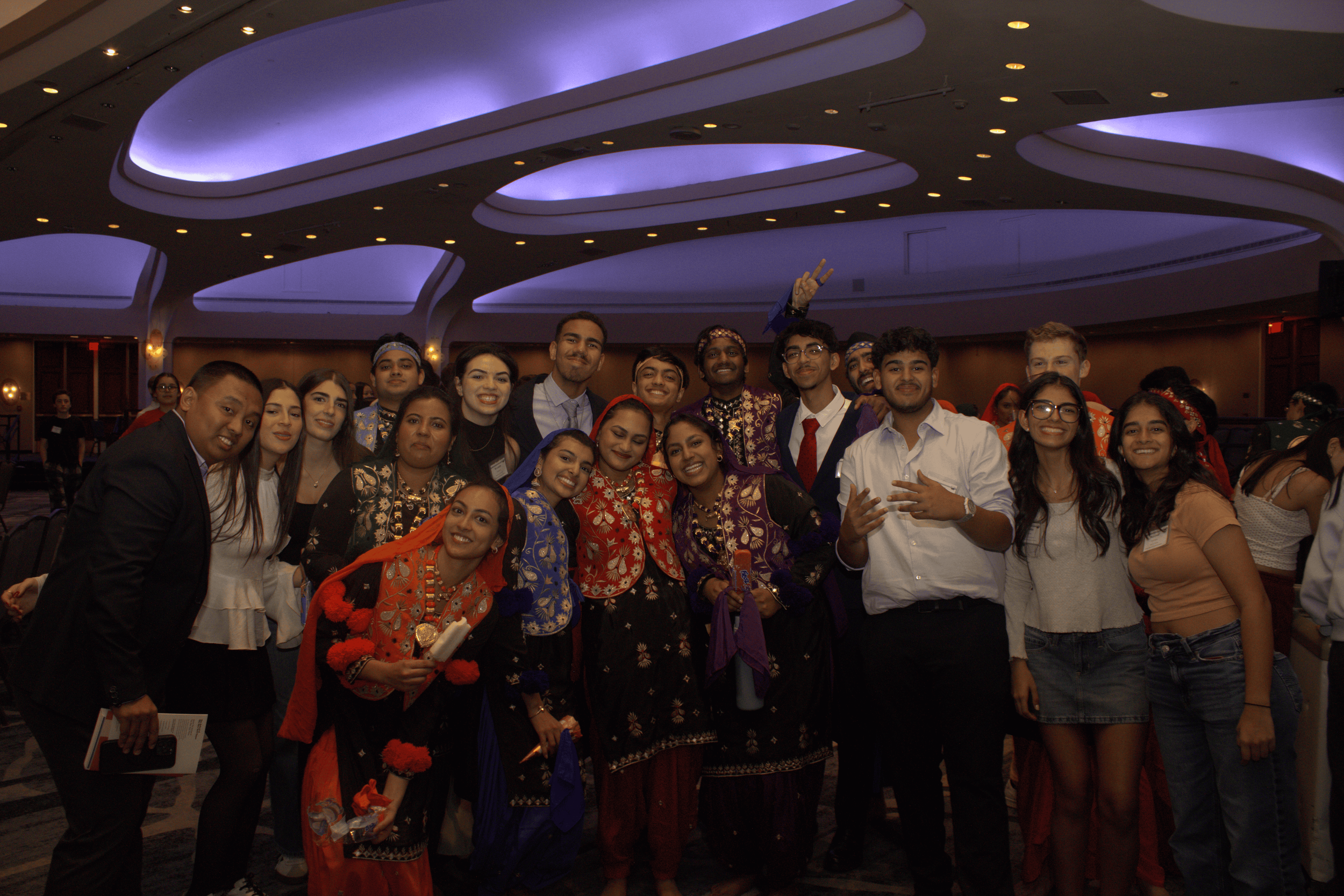 A diverse group of young adults at a formal indoor event, some wearing traditional or cultural attire, smiling and posing together.