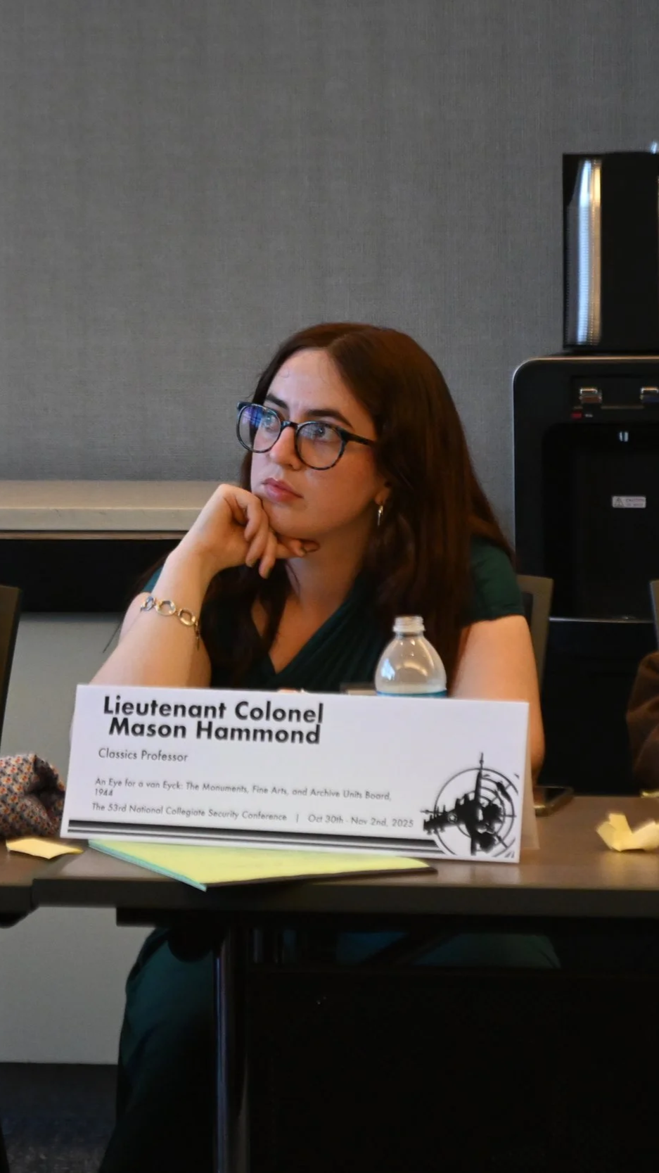 A woman with glasses sitting at a conference table, resting her chin on her hand, with a nameplate in front of her that reads 'Lieutenant Colonel Mason Hammond, Classics Professor'.