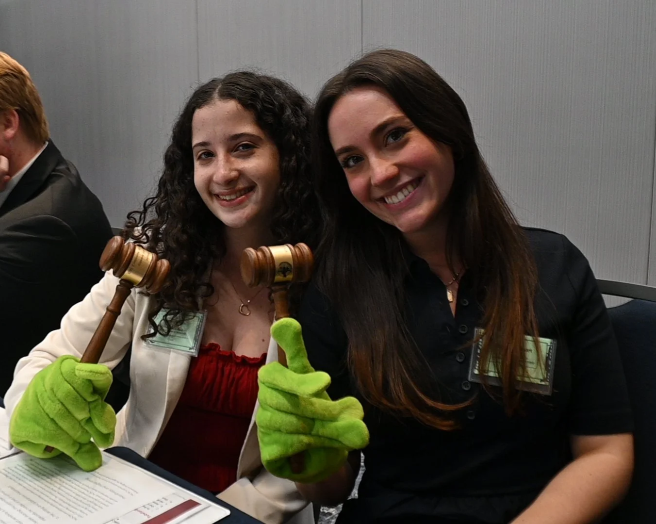 Two women smiling at a conference or meeting, holding gavel props and wearing green glove puppets, with name badges on their chests.