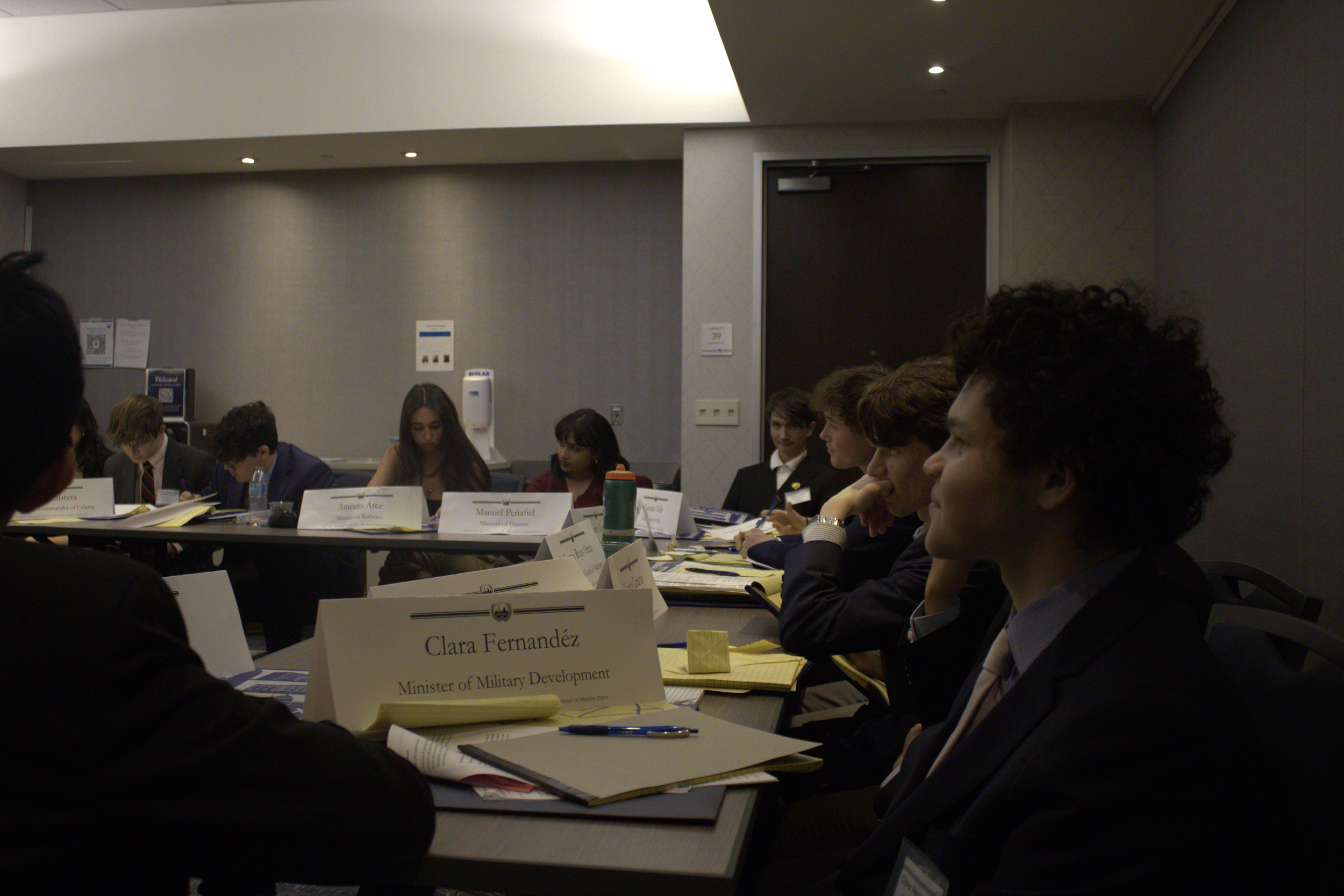 A group of young people at a formal meeting or conference, sitting around a table with name tags and documents, in a well-lit room with gray walls and a door in the background.