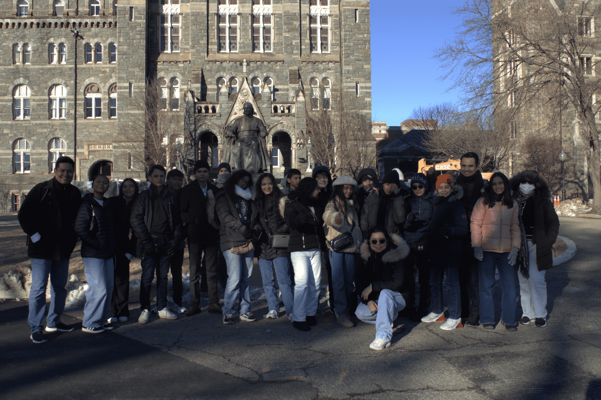 Group of diverse young people standing outdoors in front of a historic stone building with a statue of a man sitting on a chair at its center. The scene is set on a clear winter day with some snow on the ground and bare trees.