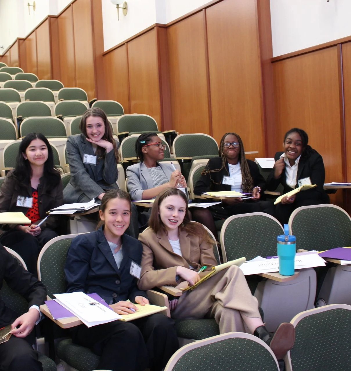 Group of diverse young women sitting in an auditorium with notebooks, pens, and a water bottle, smiling at the camera.