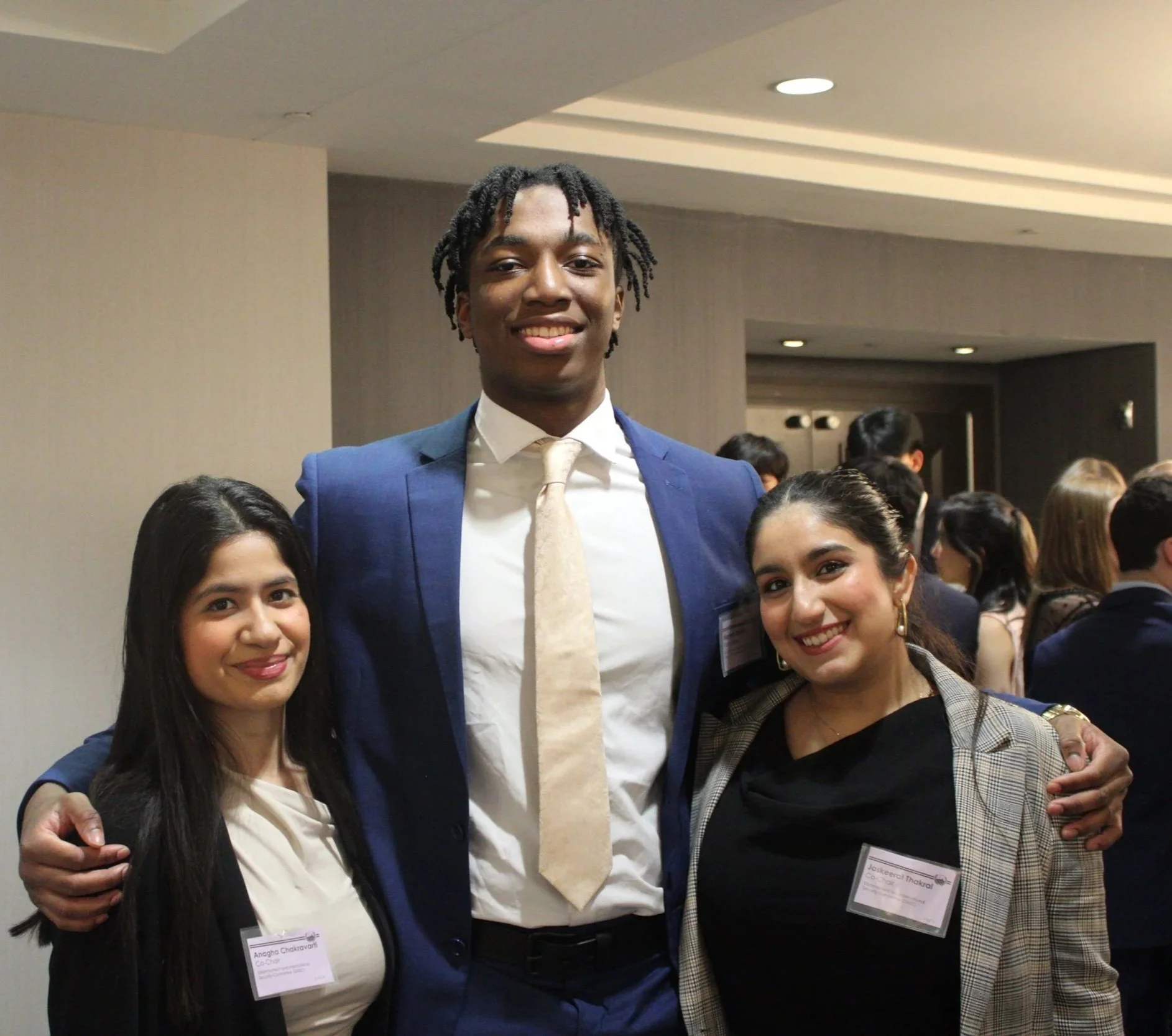 A group of diverse professionally dressed people at a social or professional event, smiling and posing for a photo. The focus is on three individuals in the foreground, with a man in a blue suit and cream tie at the center and two women on either side of him.