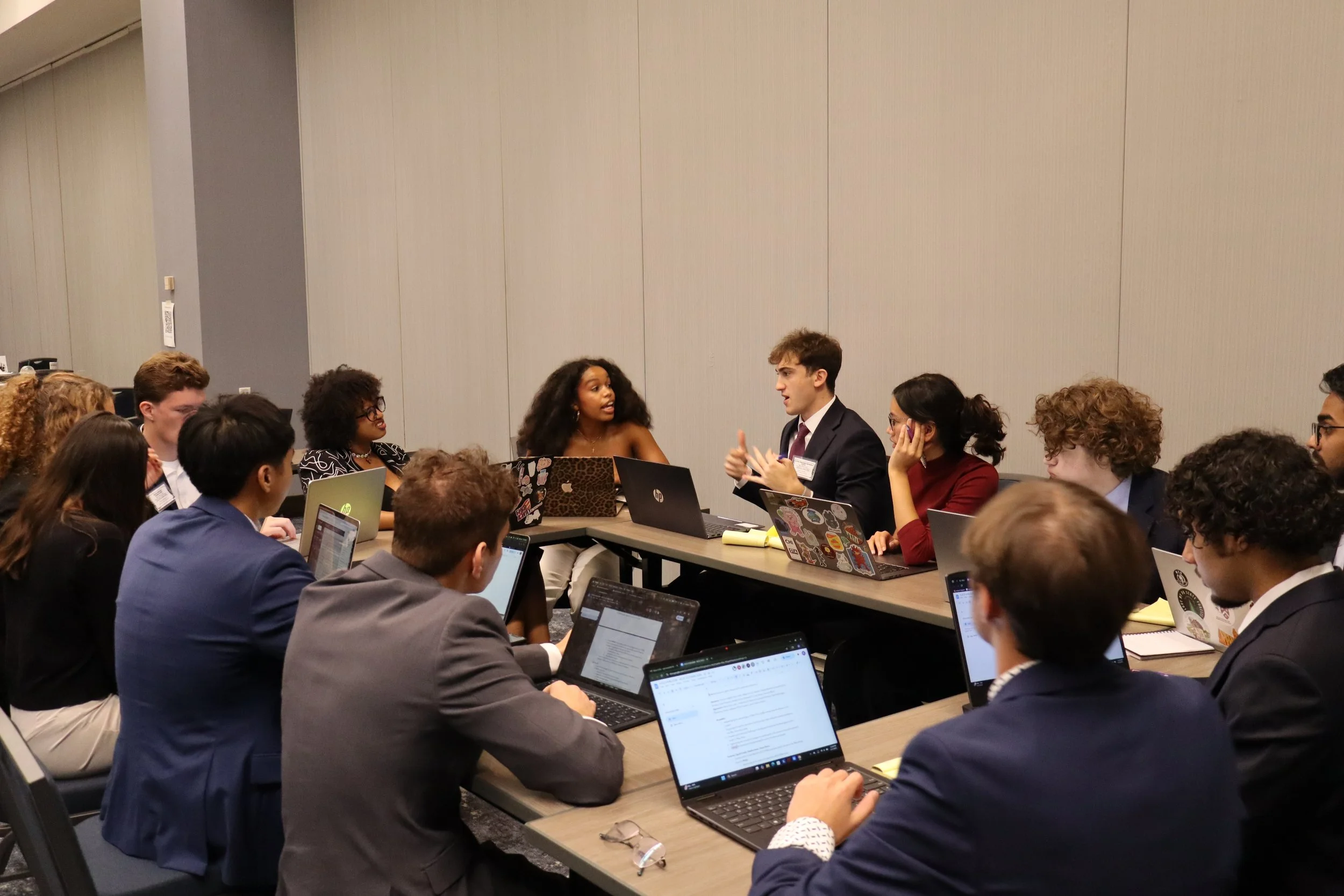 A diverse group of people gathered around a conference table in a meeting room, talking and working on laptops.