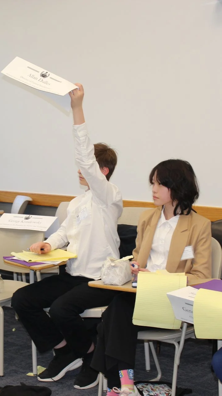 A young man and woman sitting in a classroom or conference room. The man is holding a certificate or diploma high above his head. The woman is looking to the side with papers and a notepad on her lap and a box labeled "Genius" on the table in front of her.
