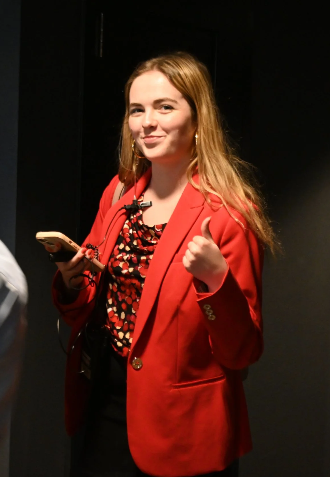 A woman with red hair, wearing a red blazer and a patterned top, smiling and giving a thumbs-up while holding a phone, standing against a dark background.