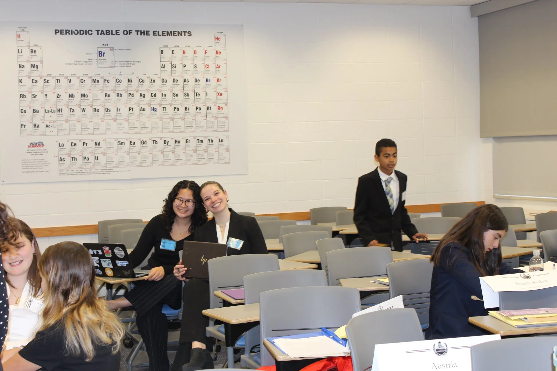 A classroom scene with several students, some working on laptops and others sitting or standing, with a large periodic table poster on the wall and a placard indicating the country Austria.