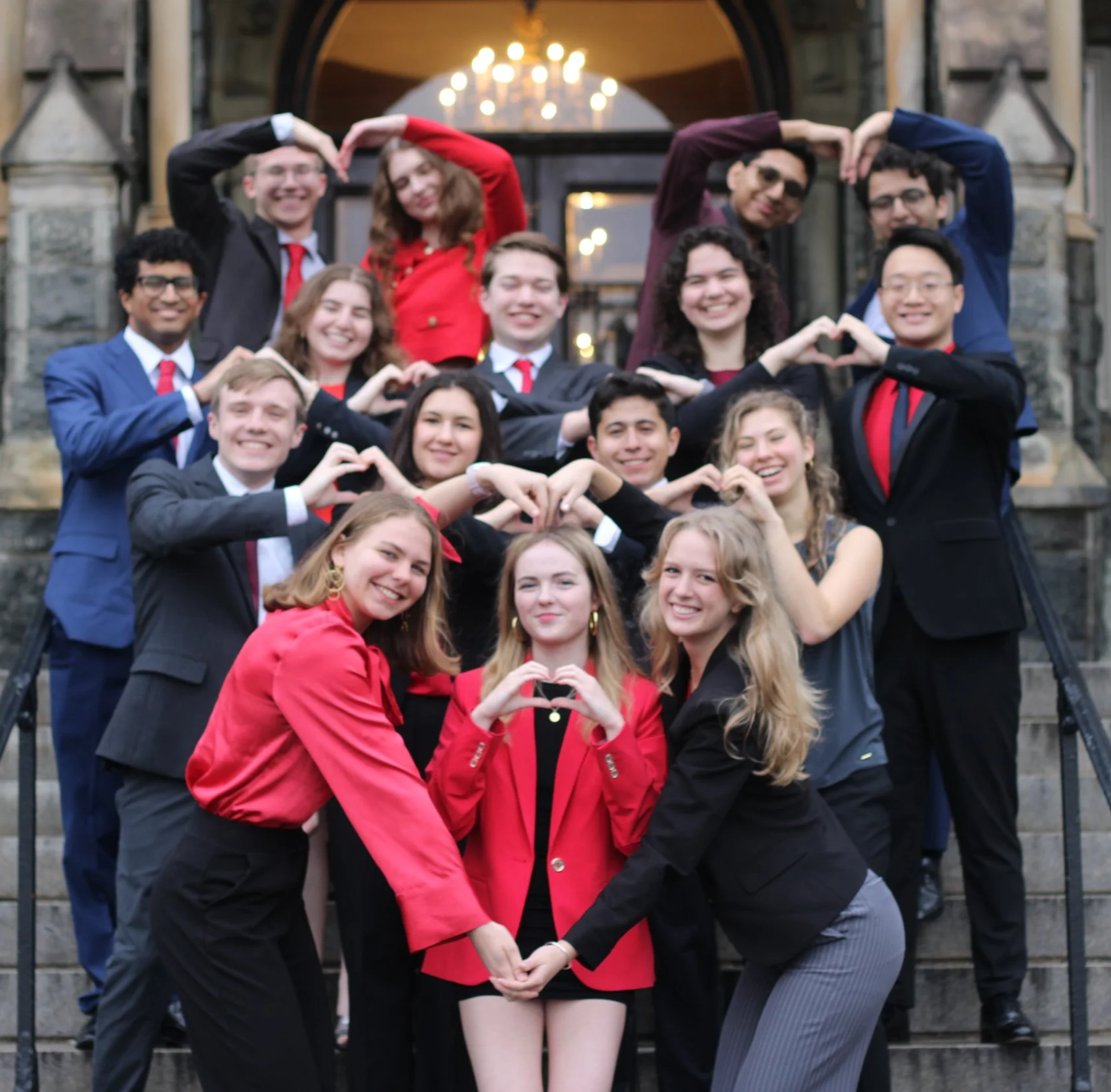 A group of young adults dressed in formal attire, smiling, and making heart shapes with their hands in front of a building with stone steps and a large window.