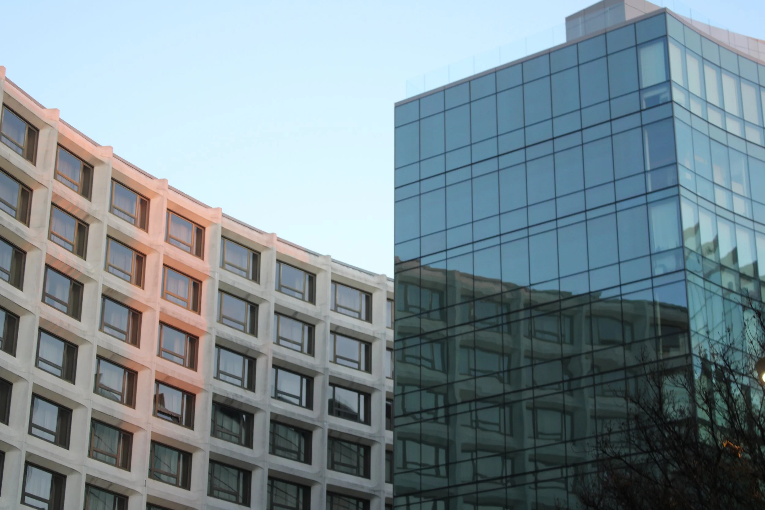 Photo of two modern buildings with different architectural styles against a clear sky. One building has a concrete facade with uniformly spaced windows, and the other is a glass skyscraper reflecting the surroundings.