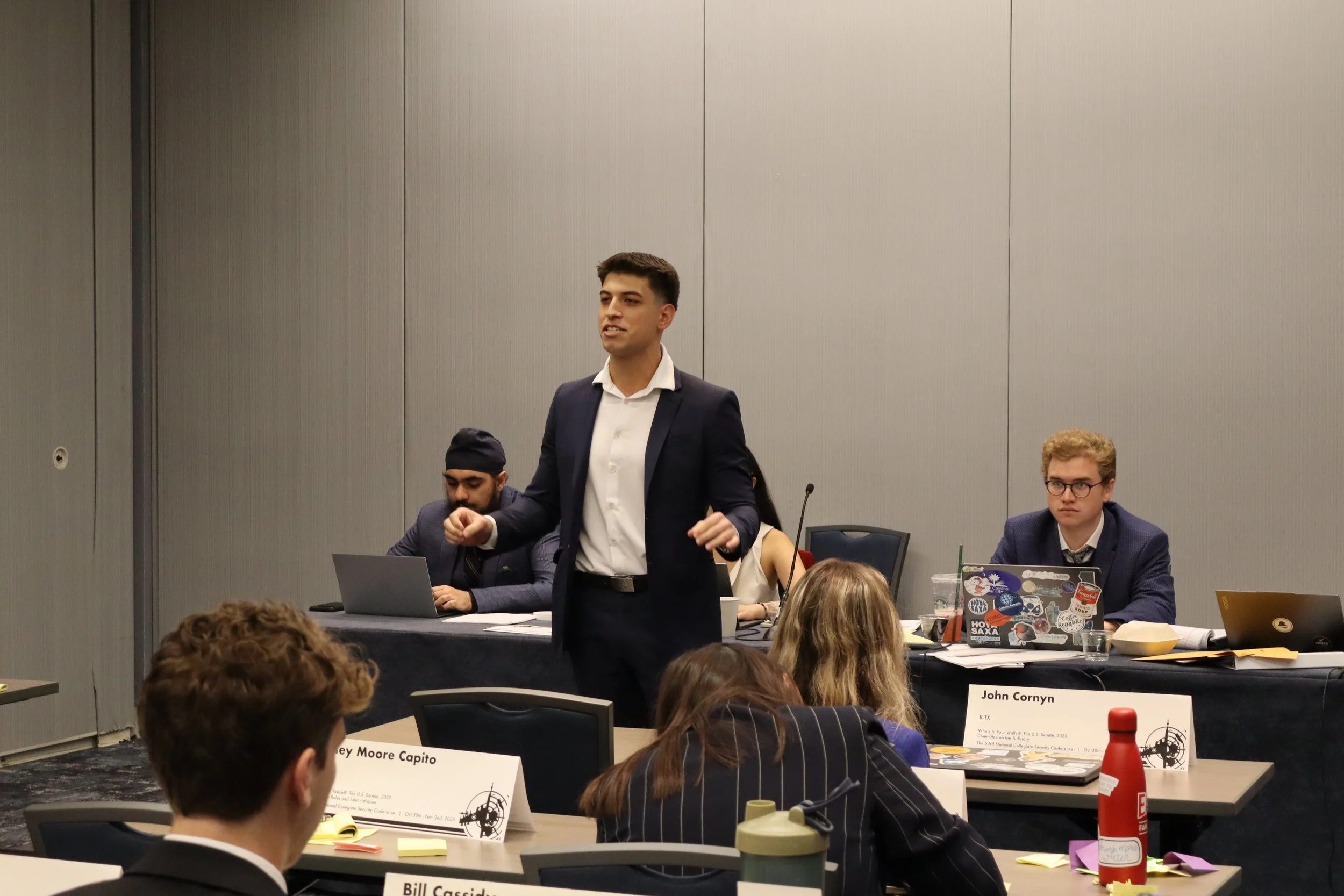 A man in a suit standing and speaking at a conference table with other seated attendees in a conference room.