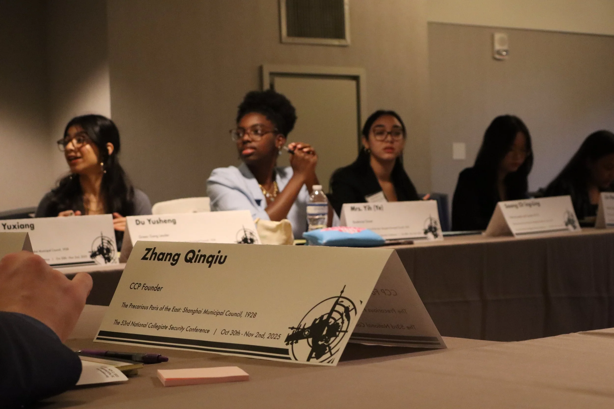 Conference attendees seated at a table, with placards visible. The focus is on a placard with the name "Zhang Qinqiu" in the foreground.