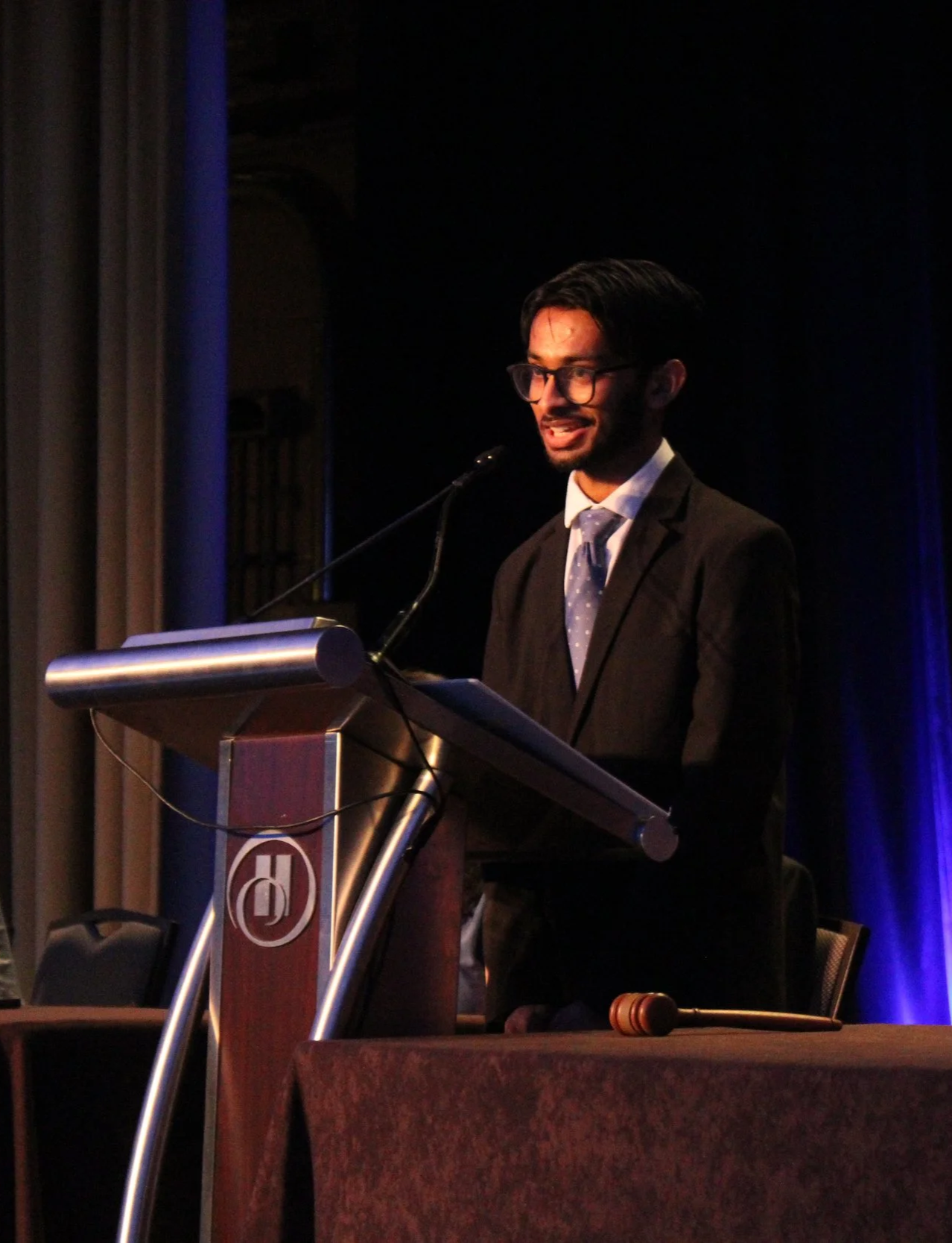 A man in a suit with glasses speaking at a podium during an event at a hotel conference room.