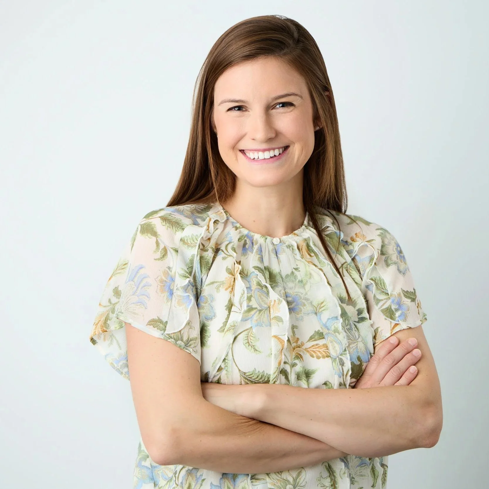 woman in short sleeve floral blouse stands with arms crossed smiling at the camera in an example of waist-up headshot framing