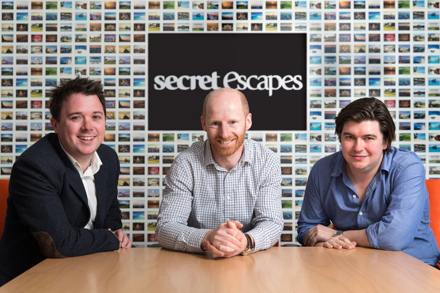 Three men sitting at a conference table in front of a wall of small photos and a large screen displaying the words "Secret Escapes."