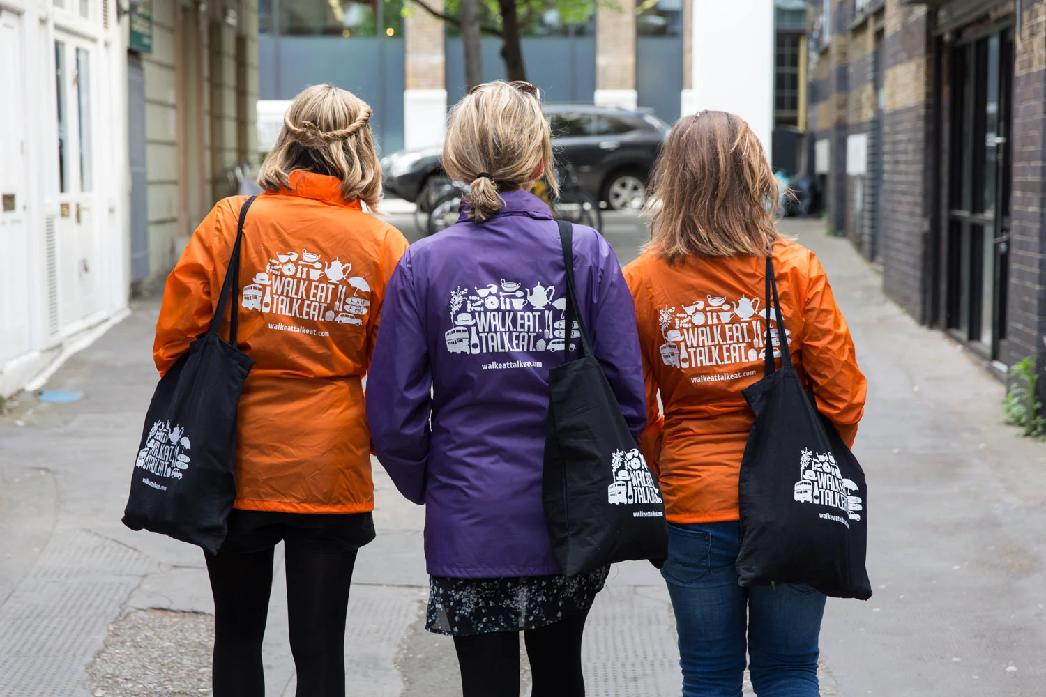 Three women walking in an alleyway, wearing jackets with a logo that says 'Walk Eat Talk Eat' and carrying black tote bags with the same logo.