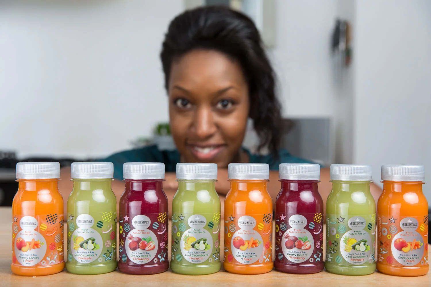 A woman looking happily at a row of colorful vegetable juice bottles on a table in a bright room.