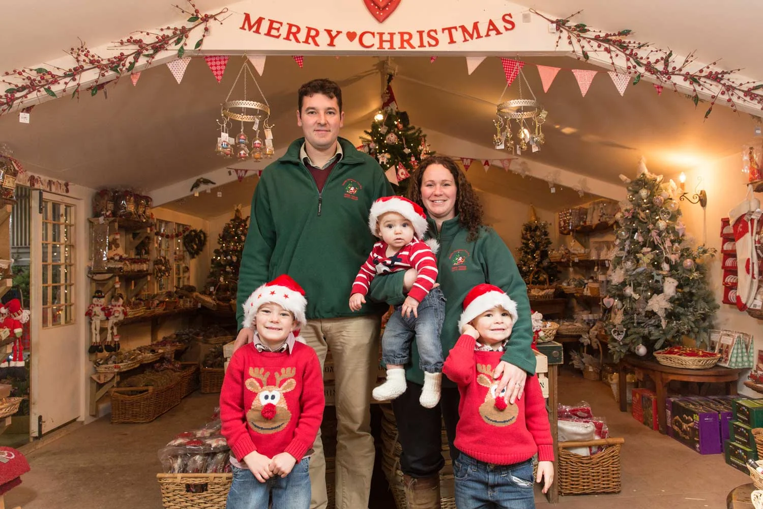 Family celebrating Christmas in a decorated store with Christmas trees, ornaments, and a "Merry Christmas" banner, wearing holiday attire including Santa hats and Christmas sweaters.