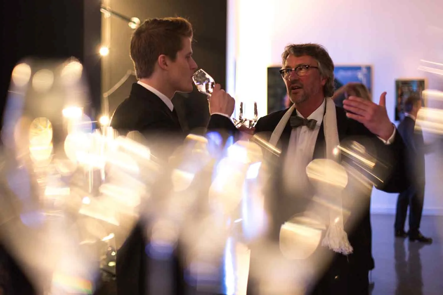 Two men in formal tuxedos and bow ties are at a fancy party, one sipping from a glass while the other gestures animatedly.