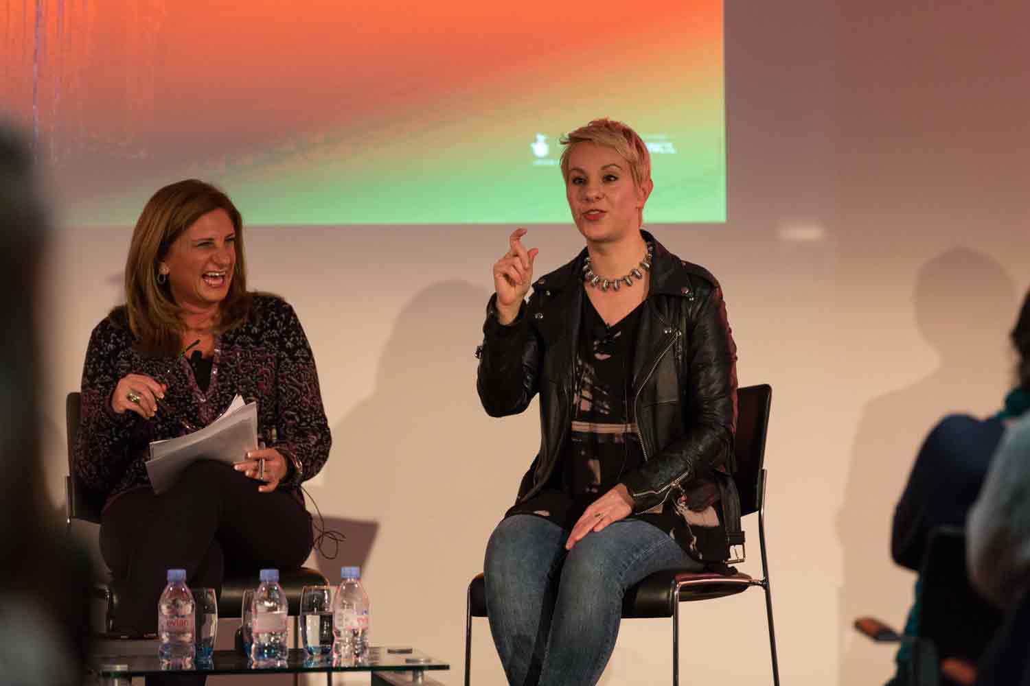 Two women sitting on chairs during a panel discussion; one woman is laughing and holding papers, the other woman is speaking and gesturing with her hand.
