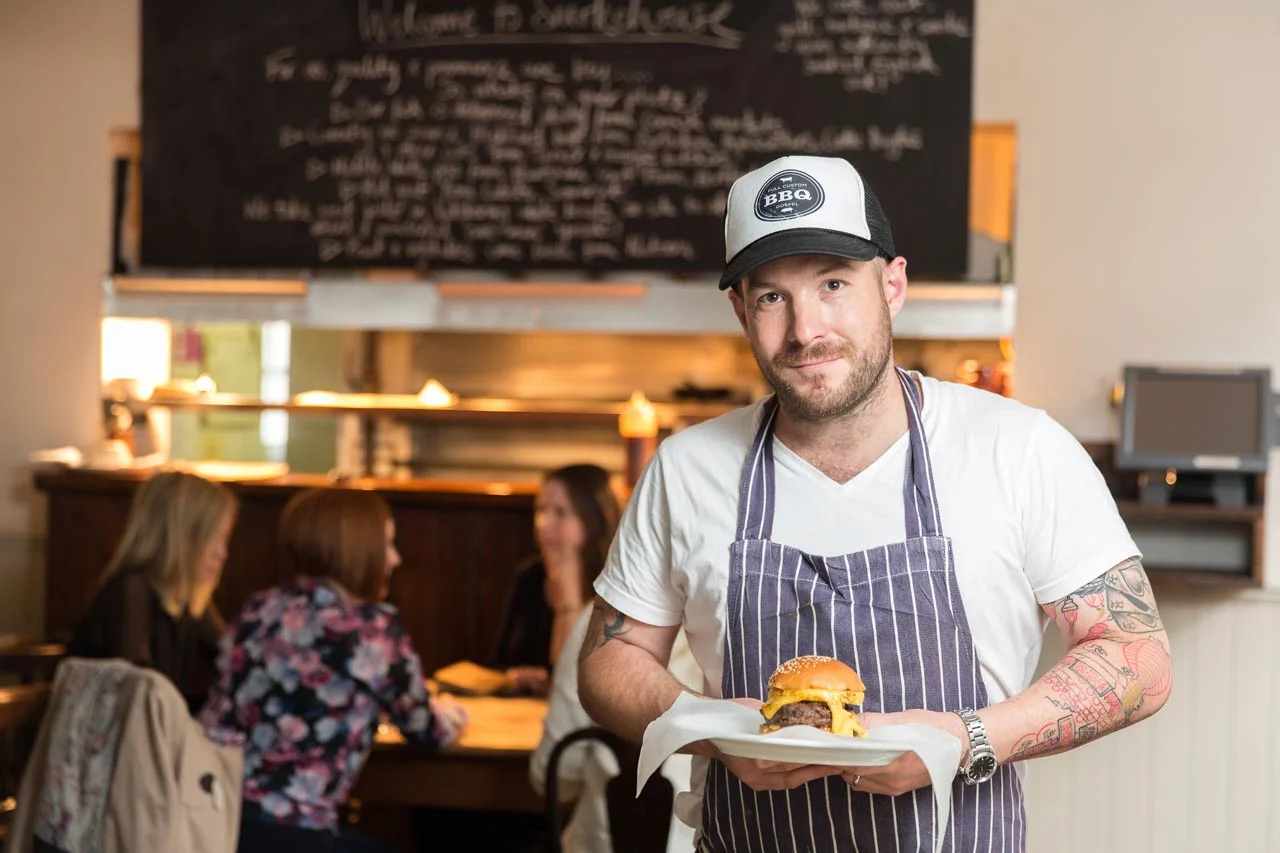A man with tattoos wearing a striped apron and a baseball cap holding a cheeseburger in a restaurant.