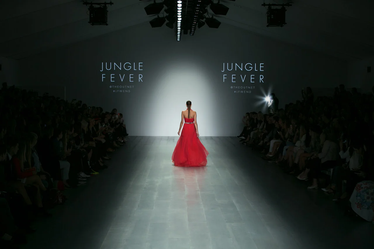 A female model in a red gown walks down a runway during a fashion show, with the audience on both sides and a bright light at the end of the runway. The backdrop displays the words 'Jungle Fever' and social media handles.