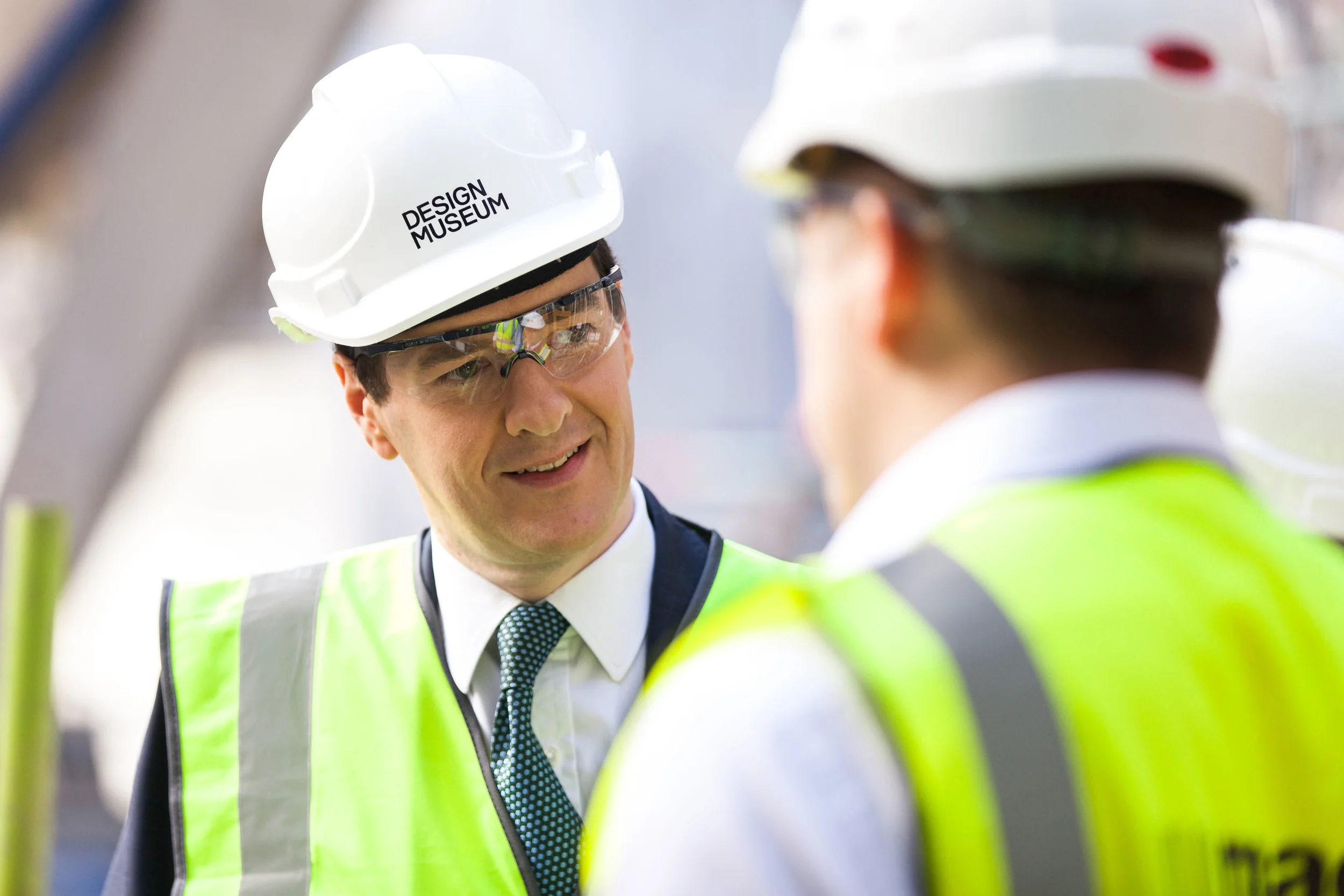 Two construction workers wearing white helmets with the words 'Design Museum' and yellow safety vests, engaged in conversation.