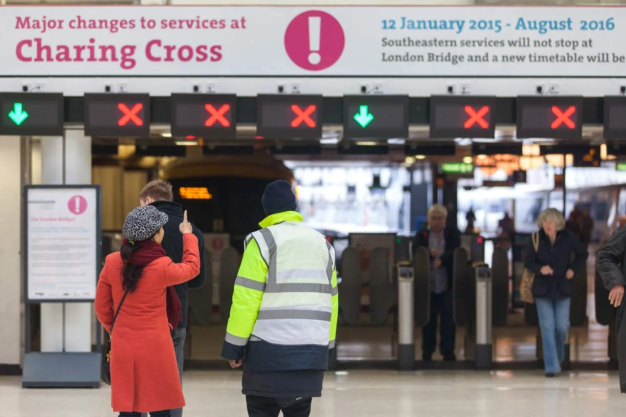 People at a train station with a sign indicating service changes. Some ticket barriers are closed with red X signals, and some are open with green arrows.