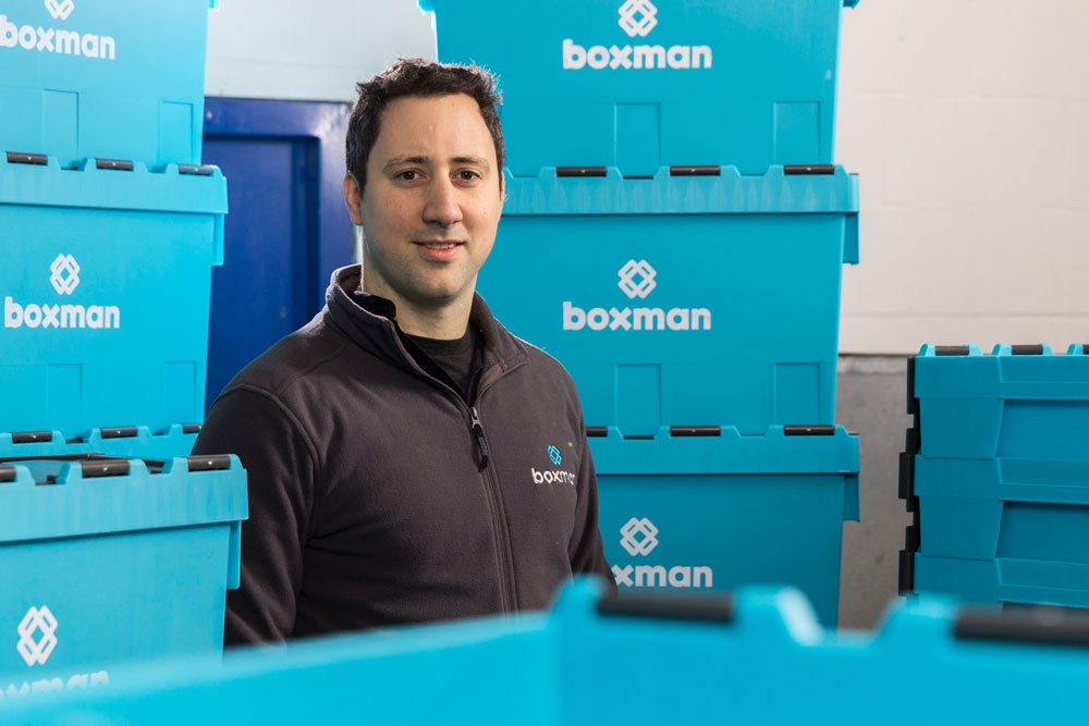 A man wearing a black zip-up jacket with a 'boxman' logo standing among large blue storage containers with matching logos in a warehouse.