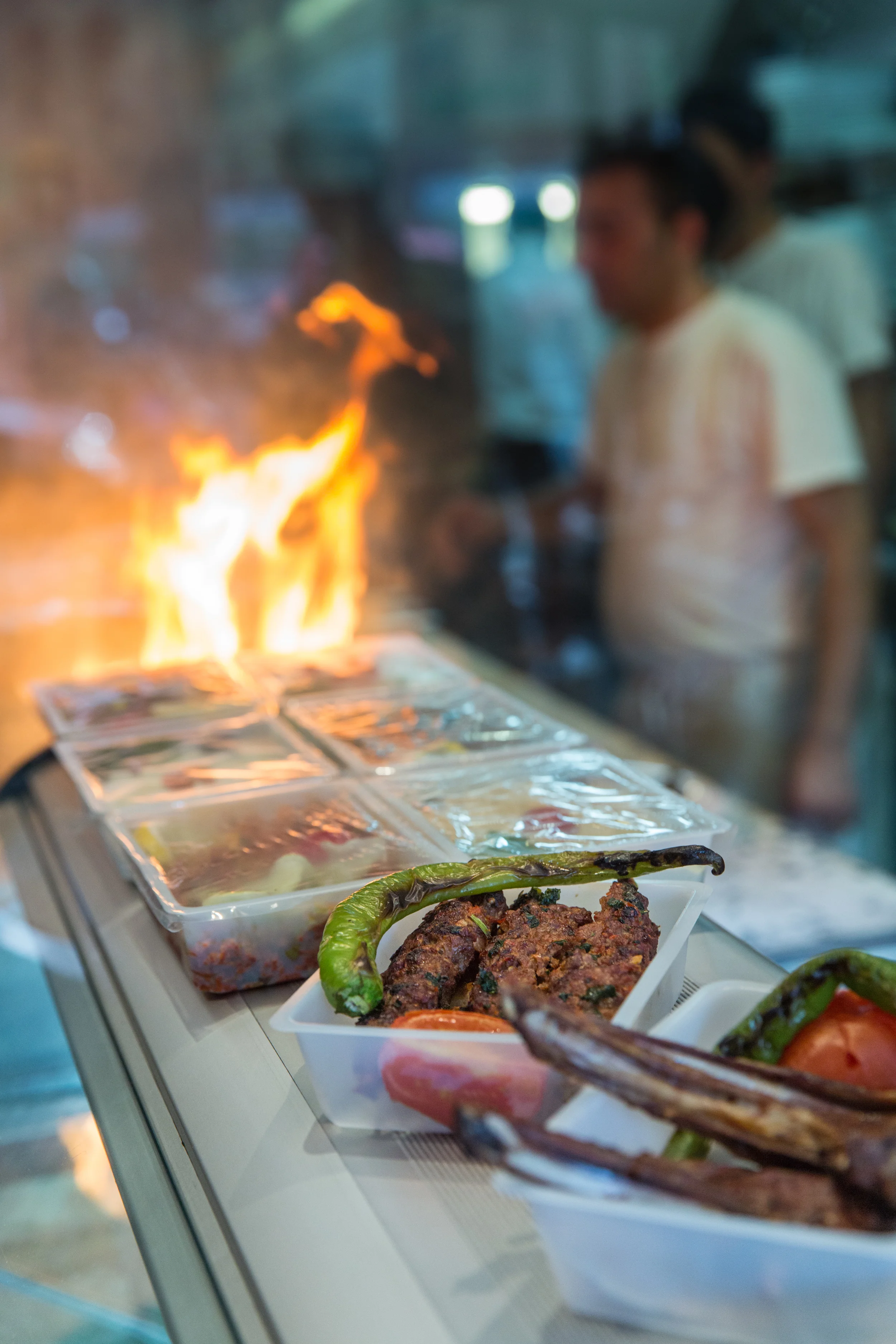 Food display at a market with grilled items, green chili peppers, and plastic containers with various ingredients, with a grill fire in the background and blurred people.