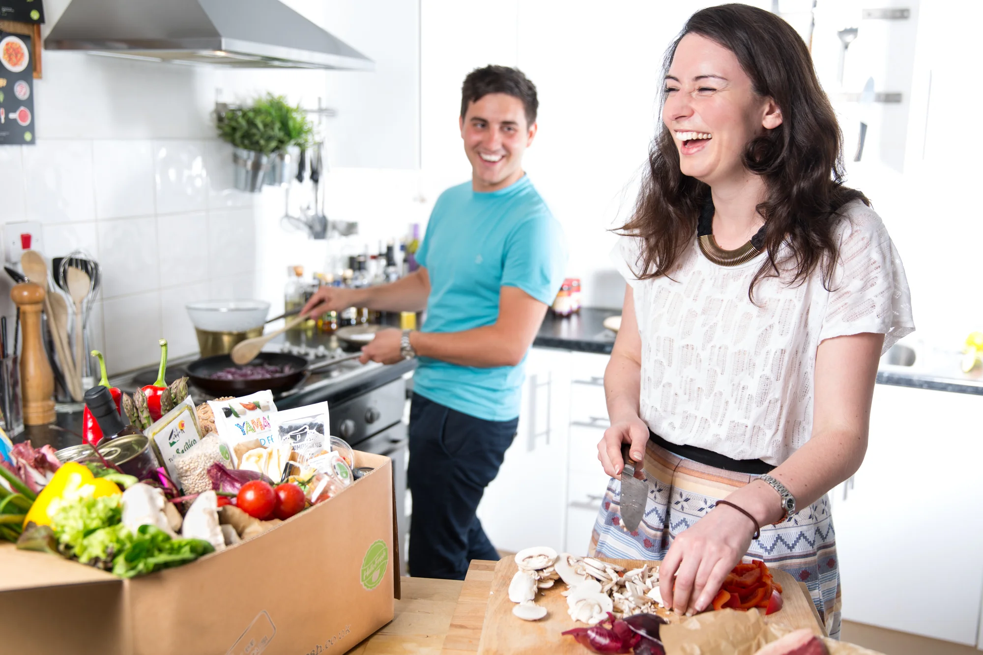 A woman is chopping vegetables on a cutting board in a bright kitchen, smiling and laughing, with a man cooking in the background.