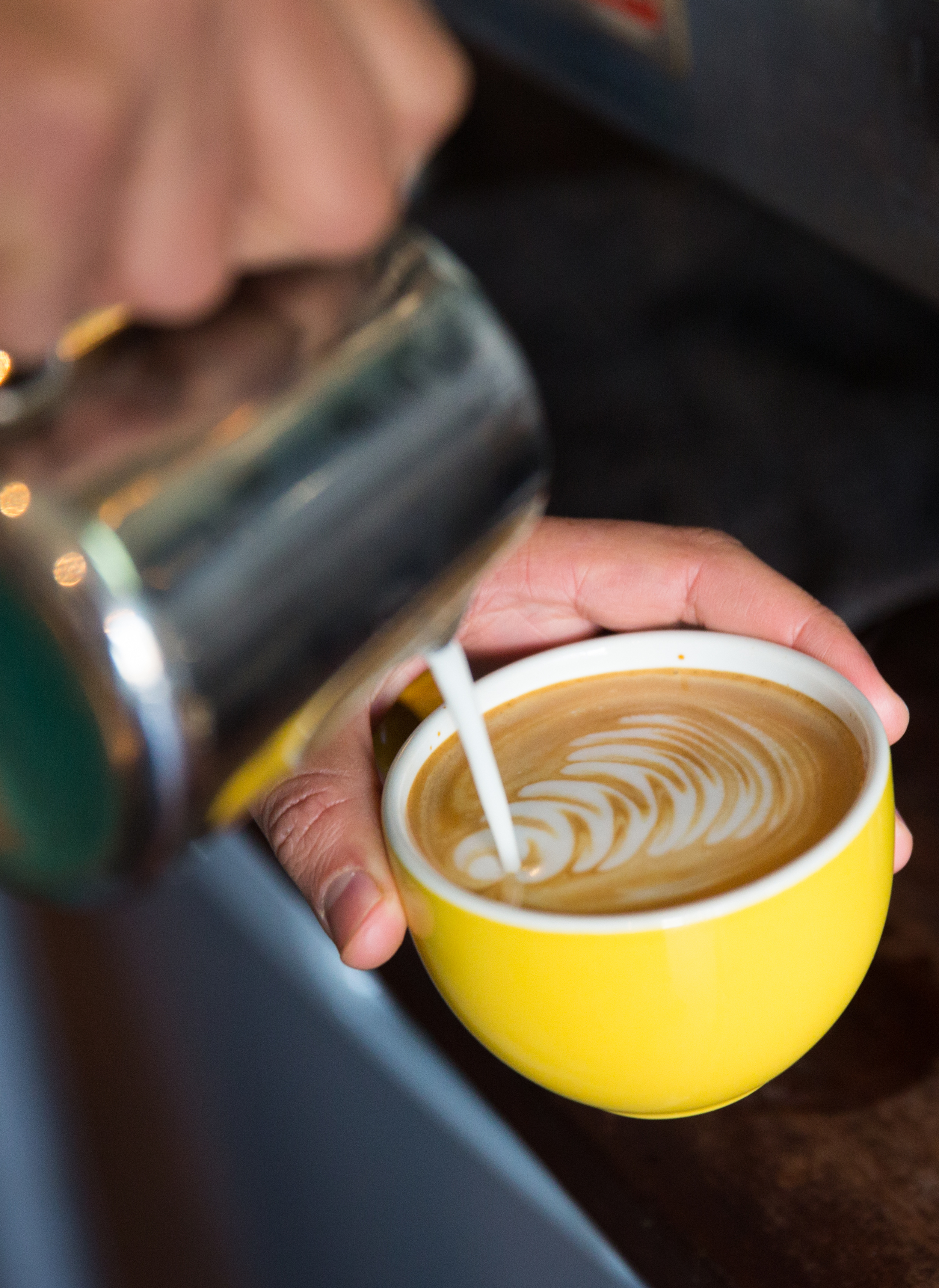 A person pouring steamed milk into a cup of coffee, creating latte art with a leaf pattern, in a yellow mug.