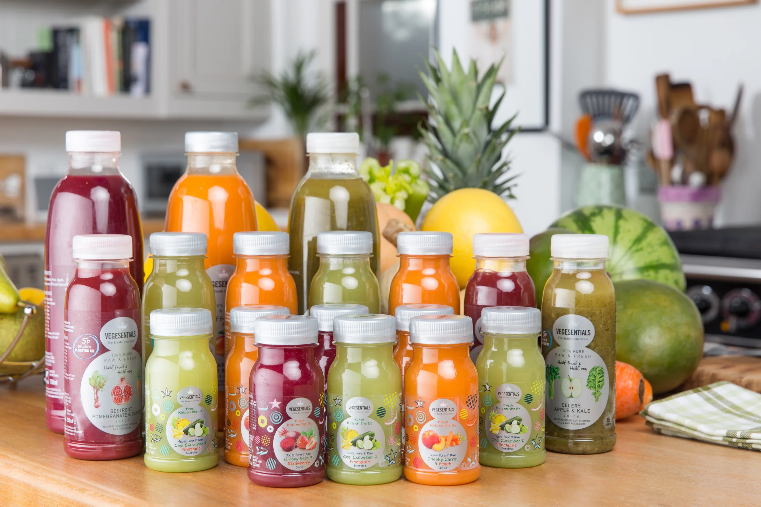 Assorted bottles of vegetable and fruit juice on a kitchen countertop with pineapples, watermelons, cantaloupe, carrots, and kitchen utensils in the background.