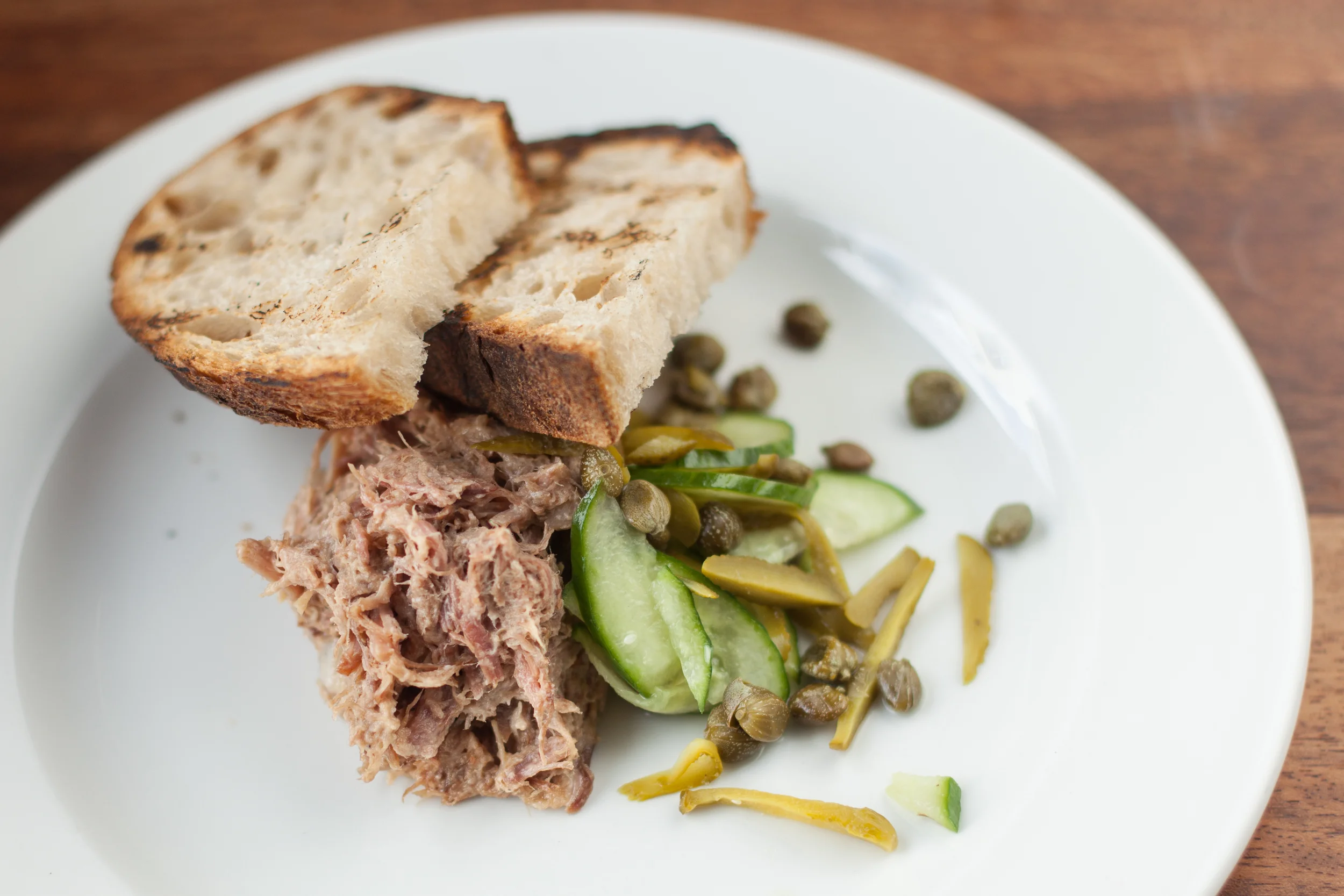 A plate with two slices of toasted bread, tuna salad, sliced cucumbers, capers, and pickled vegetables on a wooden table.