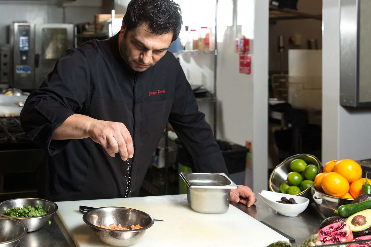 A chef sprinkling salt over a bowl of chopped vegetables in a professional kitchen.