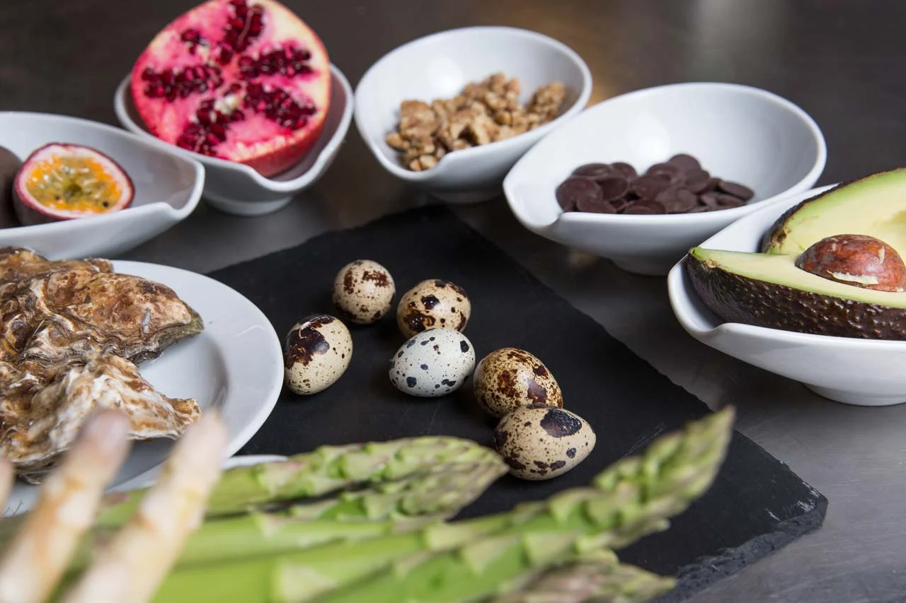 A variety of breakfast foods and healthy ingredients including quail eggs, avocado, asparagus, passion fruit, pomegranate, walnuts, and chocolate chips arranged on a black slate serving board.