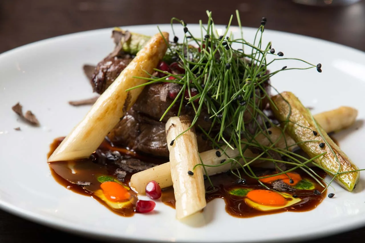 Gourmet plated dish with sliced beef, white asparagus, green vegetables, microgreens, and colorful sauces.