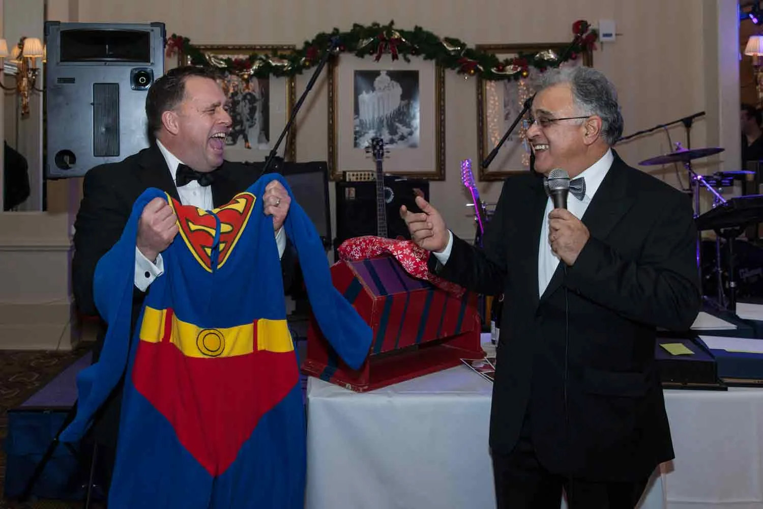 Two men in tuxedos laughing and talking at a festive event, one holding a Superman costume and the other holding a microphone, with presents and musical instruments in the background.