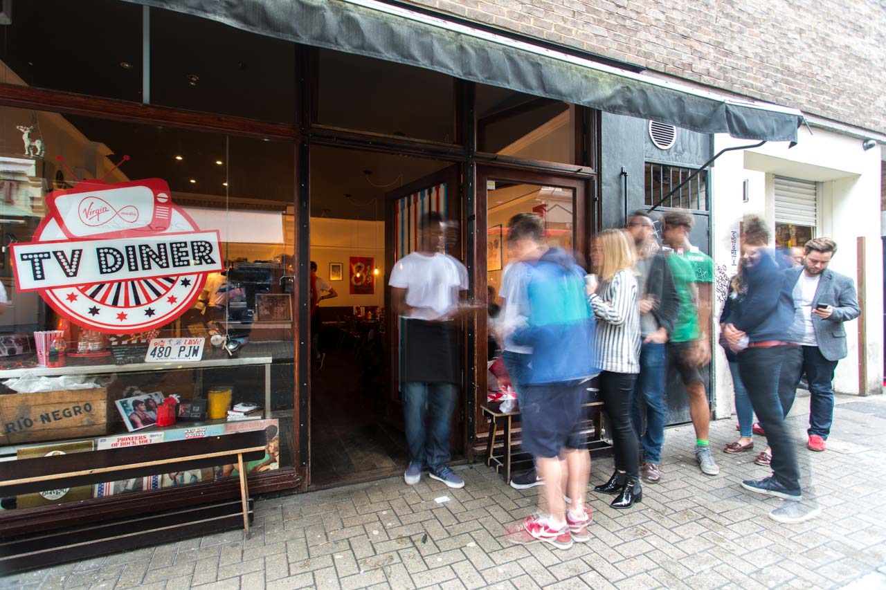 A group of people standing in line outside a restaurant called TV Diner, with some people checking their phones and others chatting, on a city sidewalk.