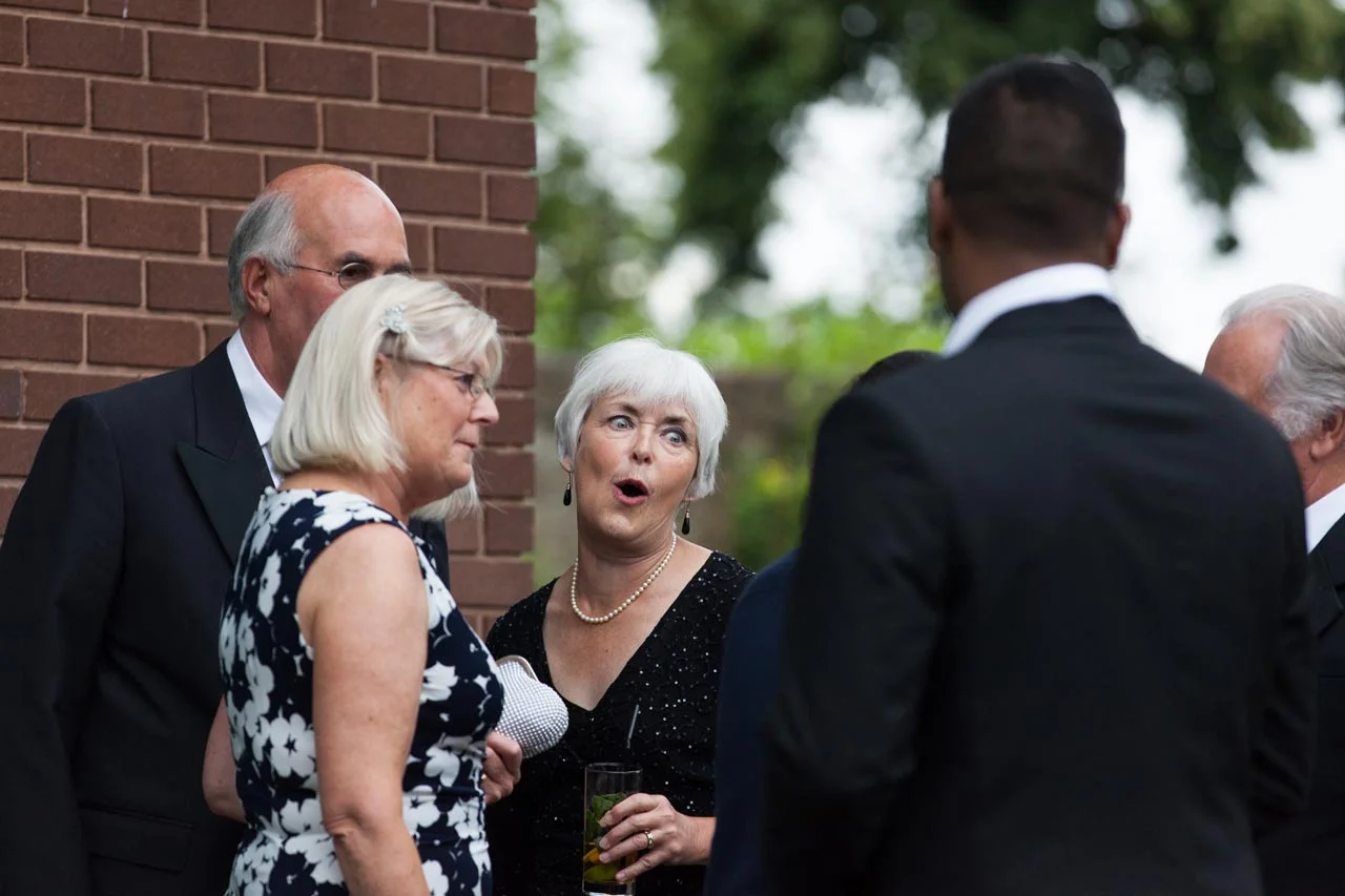 A group of six people dressed in formal attire having a conversation outside, with a brick wall and trees in the background.
