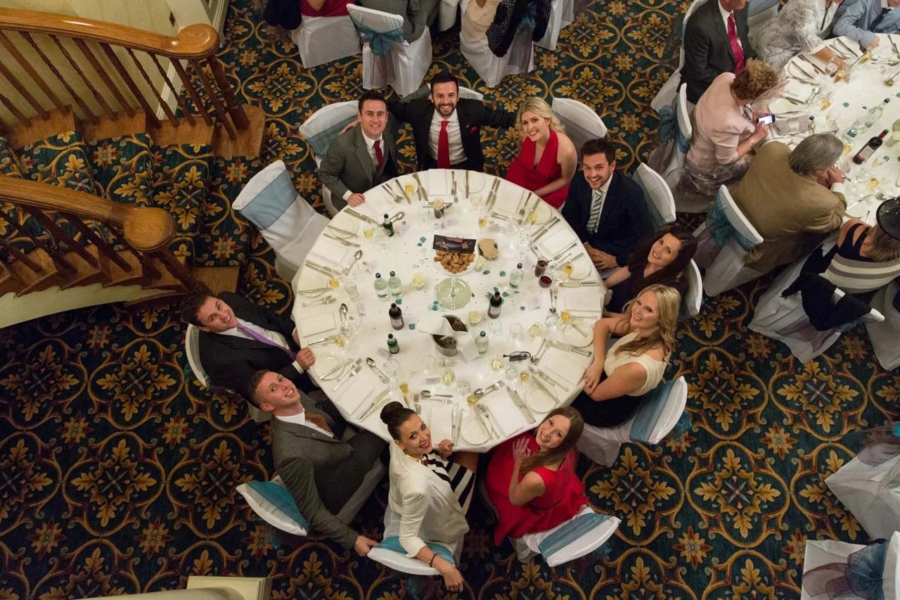 A group of people sitting around a round banquet table at a formal event, looking up and smiling at the camera.