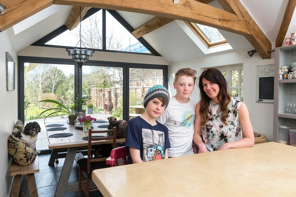 A family of three with a dog standing in a bright kitchen with vaulted ceiling and exposed wooden beams, large windows, and a view of the backyard.