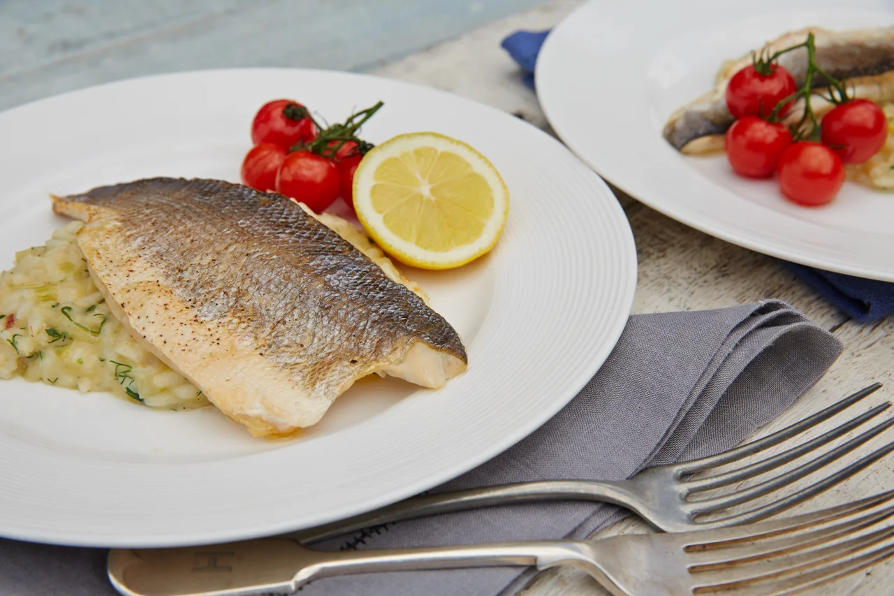 A plate with grilled fish, a lemon wedge, and cherry tomatoes. Another plate with fish and cherry tomatoes, forks, and a napkin are on a light-colored table.